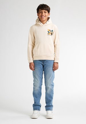 Beige hoodie met een voorzak en grafisch ontwerp in groen, oranje en bruin; gecombineerd met lichtblauwe jeans en witte sneakers.