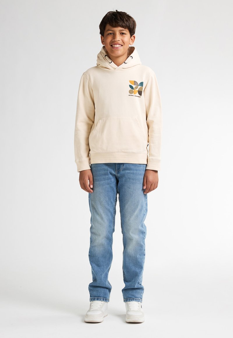 Felpa beige con tasca frontale e design grafico in verde, arancione e marrone; abbinata a jeans azzurri chiari e sneaker bianche.