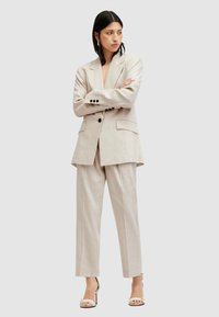 Vrouw staat met gekruiste armen in een beige oversized blazer en bijpassende broek met witte hakken op een lichte achtergrond.