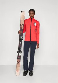 Piros cipzáras síkabát logóval, tengerészkék sínadrág és fekete síbakancsok; bézs és fekete sílécek kötésekkel a kezében; egyszerű háttér.