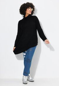 Maglione nero a maglia oversize con collo a imbuto, abbinato a jeans blu con grafiche e stivaletti argentati. Sfondo semplice.