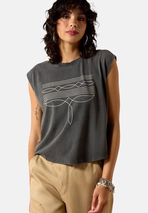 Frau mit lockigem, dunklem Haar, die ein ärmelloses dunkelgraues Oberteil mit weißem geometrischem Stichmuster und beige Hose trägt, accessoriert mit Armbändern.