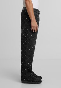 Sānu skats uz personu, kura valkā melnas džinsus ar dimanta rakstu, kas izrotāts ar mazām baltām kniedēm, un melnām kedām uz vienkārša fona.