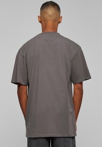 Homme avec les cheveux courts portant un t-shirt ample et uni de couleur gris foncé et un pantalon sombre, vu de dos sur un fond gris uni.