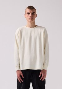 Long-sleeve crewneck shirt in light cream, featuring a textured waffle pattern, raglan sleeves, and ribbed cuffs, paired with dark pants.