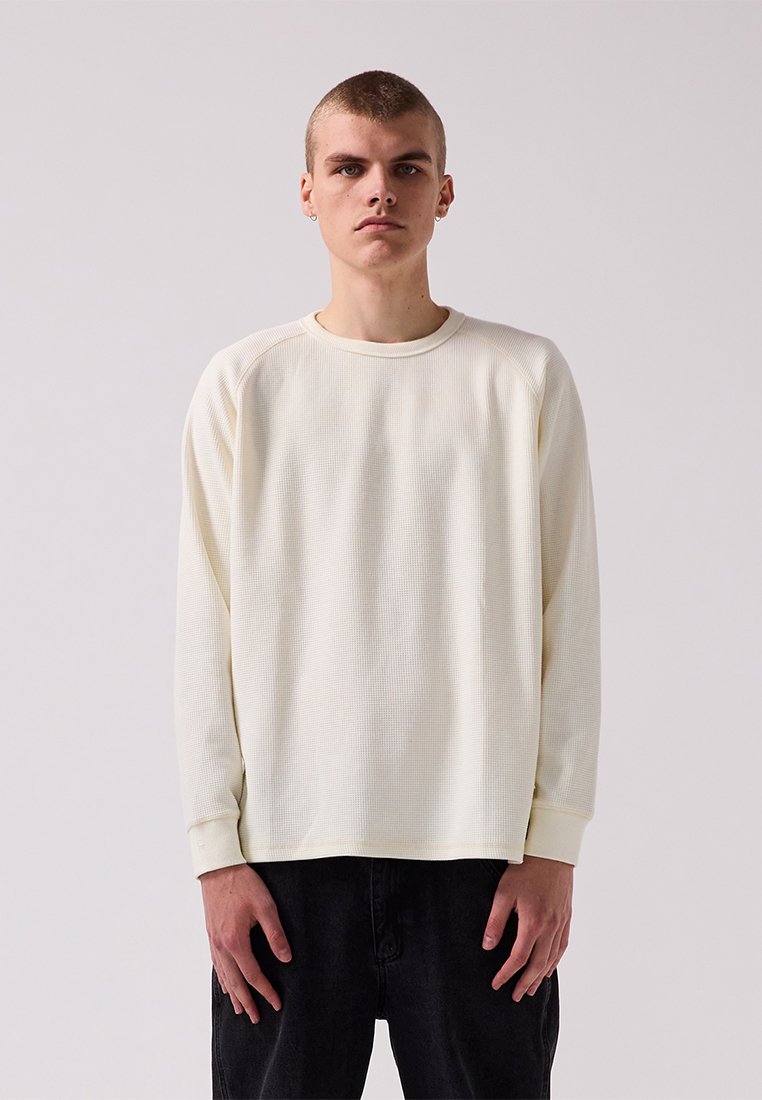 Pitkähihaisessa crewneck-paidassa on vaalea kerma väri, jossa on teksturoitu vohvelikuvio, raglanhihat ja ribatut hihansuut, yhdistettynä tummiin housuihin.