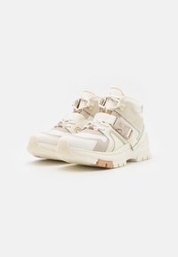 Ljusbeige sportskor med en ovandel av blandat material, rem och snörning, texturerad sula och en svag hint av persika på sulans kant.