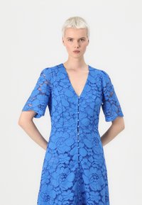 Blue lace dress featuring a deep V-neck, short puff sleeves, and decorative button closure down the front. Floral pattern throughout.