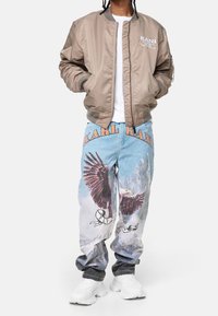 Khaki bomberjakke med ribstrikkede manchetter, hvid t-shirt og denimjeans med ørn-grafik og teksten "KARL" i orange, kombineret med hvide sneakers.