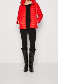 Chaqueta acolchada roja con costuras horizontales y capucha, combinada con leggings negros ajustados y botas altas de cuero negro hasta la rodilla.