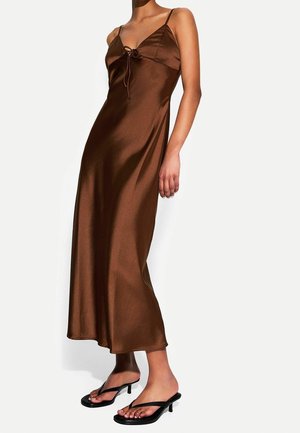 Brown satin maxi dress with a V-neckline, adjustable straps, and a bow accent at the front; paired with black heeled sandals.