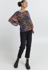 Blusa trasparente con motivo floreale colorato in rosa, blu e arancione, dotata di maniche larghe e scollatura rotonda. Indossata con pantaloni neri e stivaletti.