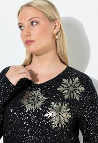 Černý svetr s výstřihem do V a metalickými vzory sněhových vloček. Látka má jemnou tečkovanou strukturu, která umocňuje celkový design.