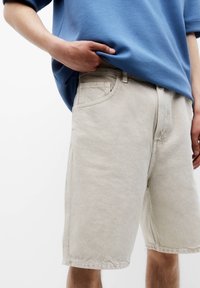 Persona con pantalones cortos de mezclilla beige y una camiseta azul de gran tamaño, con la mano metida debajo de la camiseta cerca de la cintura.