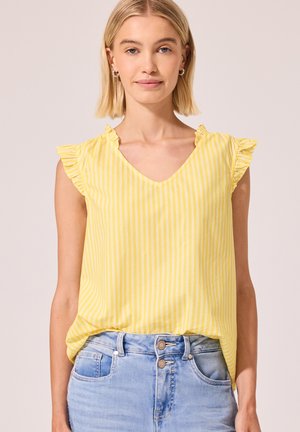 Kvinde med kort blondt hår iført en gul ærmeløs top med striber og flæser på skuldrene samt lyseblå jeans med dobbelte knapper.