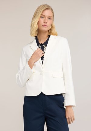 Kobieta o blond włosach nosi biały, dopasowany żakiet na czarnej, wzorzystej koszuli oraz granatowych spodniach, stojąc na tle jednolitego beżowego tła.