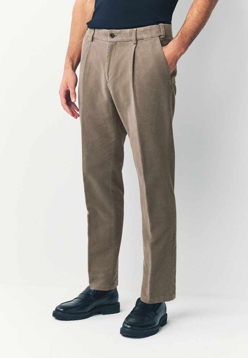 Pantalon chino en tissu marron clair à texture douce, avec des plis à l'avant, des poches latérales et une fermeture par bouton à la taille.