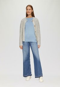 Grijze gebreide cardigan met houten knopen, lichtblauwe trui en wijde blauwe jeans, gecombineerd met witte sneakers; neutrale achtergrond.