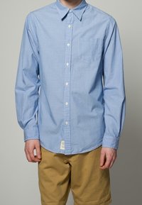 Sininen raidallinen nappipaita, jossa on kaulus, pitkät hihat ja rintatasku. Sileä kangas, valkoiset napit ja alareunassa oleva etiketti.