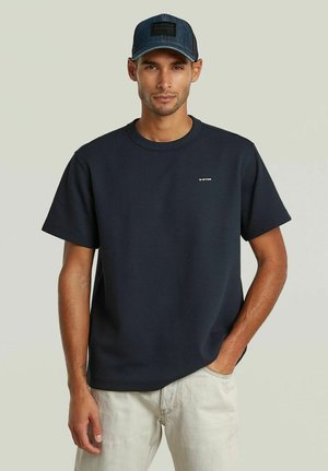 Uomo con camicia blu navy a maniche corte con logo "G-STAR" e cappellino da baseball blu scuro, pantaloni beige, in piedi con una mano in tasca.