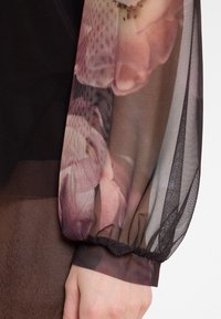 Durchsichtiger schwarzer Netzärmel mit pinkem Blumenmuster und gerafftem Bündchen über nacktem Arm und Hand.