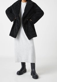 Manteau noir texturé avec col à revers, porté sur une robe côtelée gris clair. Des bottes noires à semelles épaisses complètent le look.