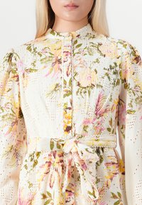 Donna in un vestito crema con fantasia floreale, maniche a sbuffo, bottoni frontali, dettagli in pizzo e una cintura annodata in vita.