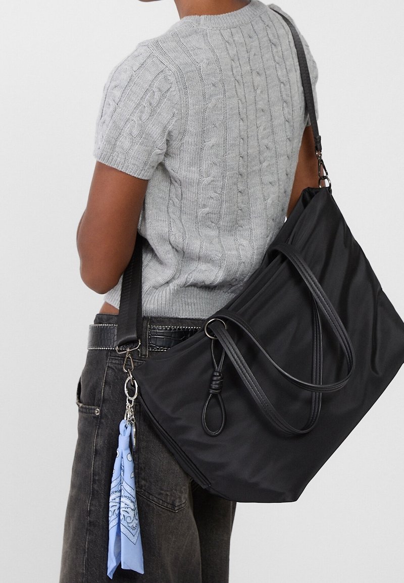 Personne portant un pull gris à manches courtes en maille torsadée avec un jean foncé et portant un grand sac à bandoulière noir avec un porte-clés bandana bleu.