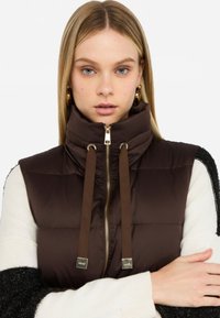 Gilet imbottito marrone con colletto alto, cerniera dorata e coulisse marroni. Indossato sopra un top a maniche lunghe bianco e nero con maniche texturizzate.