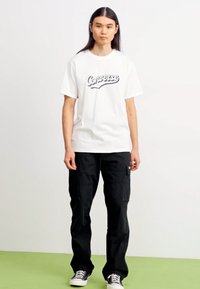 Hvit bomulls t-skjorte med svart "Converse"-logo, kombinert med svarte cargo-bukser og svart-hvite joggesko, stående på et grønt gulv.