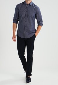 Mężczyzna w niebieskiej, długiej koszuli z guzikami, z podwiniętymi rękawami, czarnych spodniach i granatowych trampkach, idzie z jedną ręką w kieszeni.