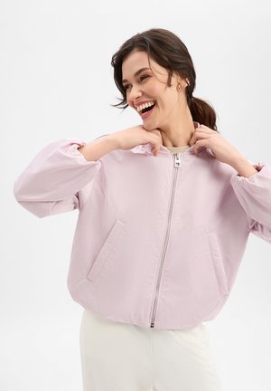 Kobieta uśmiecha się i poprawia kołnierz jasnoróżowej kurtki na zamek z kieszeniami, ubrana w białe spodnie, na jednolitym tle.