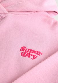 Felpa rosa realizzata in tessuto morbido con polsini a coste, caratterizzata da un logo ricamato "Super Dry" di colore rosa scuro sul davanti.