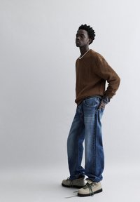 Bruine corduroy trui, loszittende blauwe jeans, grijs gelaagd shirt eronder. Textured beige schoenen met groene veters, staand tegen een grijze achtergrond.