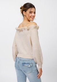 Beige off-the-shoulder sweater with ruffled edge and ribbed cuffs, paired with light blue fitted jeans, showcasing a soft texture.