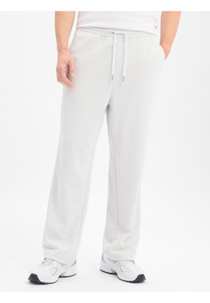 Personne portant un pantalon de survêtement blanc à cordon de serrage et des baskets blanches, debout, les mains partiellement dans les poches, sur un fond blanc.