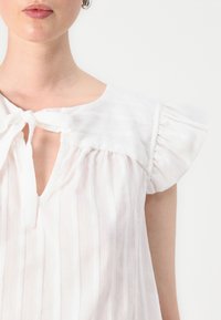 Blusa bianca con maniche a sbuffo, dettagli con cravatta davanti e righe verticali. Realizzata in tessuto leggero con una vestibilità comoda.