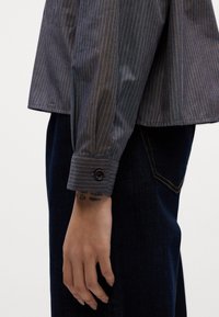 Donkerblauw shirt met lange mouwen en dunne oranje strepen, voorzien van een knoop op de manchetten. Gecombineerd met een donkere spijkerbroek.