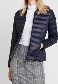 Doudoune bleu marine avec un design matelassé, col montant et poches zippées, associée à une jupe à motifs avec des accents en niveaux de gris et en bleu.