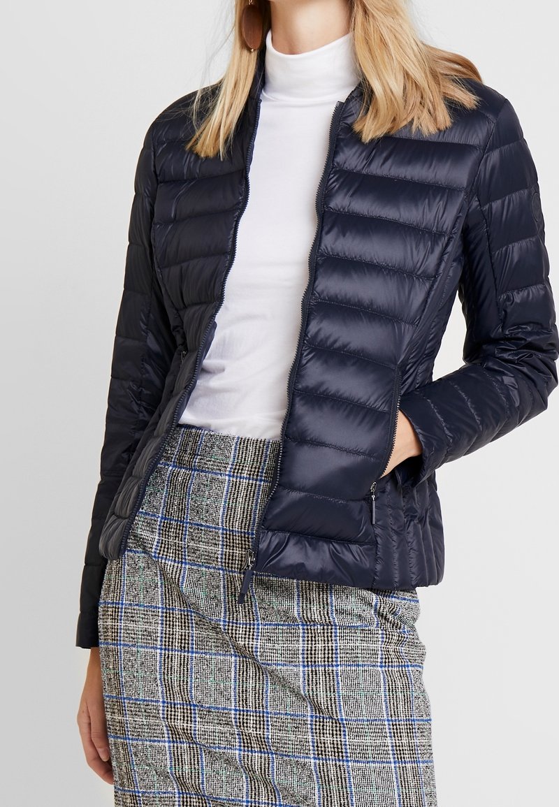 Doudoune bleu marine avec un design matelassé, col montant et poches zippées, associée à une jupe à motifs avec des accents en niveaux de gris et en bleu.