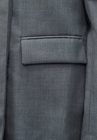 Γκρι σακάκι από ύφασμα με λεία υφή, που διαθέτει πορτοφόλι στον αριστερό πλευρό, ραμμένο σχέδιο και διακριτικό διαγώνιο μοτίβο ύφανσης.
