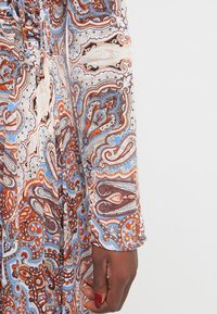 Chemisier à boutons en tissu léger avec un motif paisley multicolore. Présente des teintes de bleu, orange et marron avec des détails froncés aux manches.