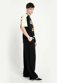 Młody mężczyzna w czarnych szerokich spodniach, koszulce z grafika w kolorze czarnym i beżowym, czarnych trampkach oraz srebrnej bransoletce, stoi bokiem.