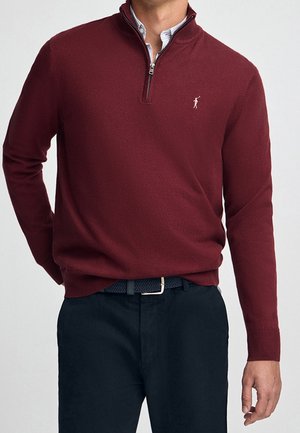 Homme portant un pull bordeaux à fermeture éclair sur une chemise à col et un pantalon noir avec une ceinture tissée noire, debout avec une main le long du corps.