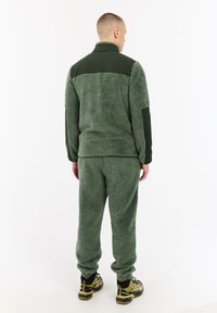 Hombre de pie de espaldas, vistiendo una chaqueta y pantalones de forro polar verdes, con zapatos de senderismo, sobre un fondo blanco.