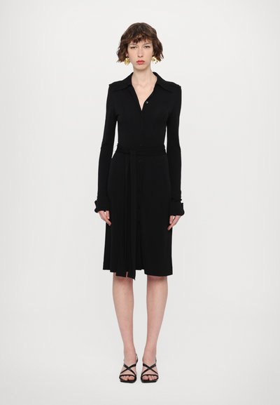 Čierne šaty s dlhým rukávom, so zapínaním na gombíky vpredu, golierom, opaskom na viazanie v páse a rozšíreným lemom. Hladká látka bez viditeľných vzorov alebo textúr.