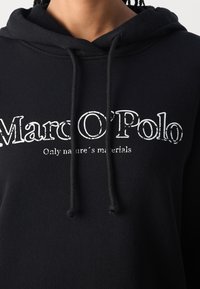 Sudadera con capucha negra hecha de tela suave, con un bolsillo canguro frontal, capucha con cordón y texto en blanco que dice "Marc O’Polo Solo materiales naturales."