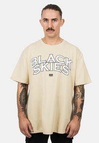 Krém színű, oversized póló, amelynek elülső részén a "BLACK SKIES" felirat található kiemelkedő fekete körvonalban. Rövid ujjú, sima anyagú, látható tetoválásokkal a karokon.