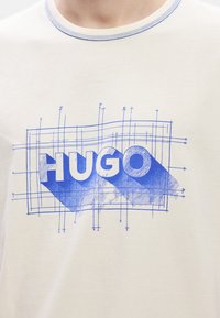 T-shirt de algodão em bege claro com um estampado gráfico azul que apresenta a palavra "HUGO" e elementos geométricos em linha. Costura de contraste na gola.