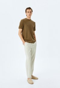 Braunes strukturiertes T-Shirt, hellfarbene Leinenhose und geflochtene Slipper. Das Modell steht gerade mit den Händen in den Taschen vor einem hellen Hintergrund.